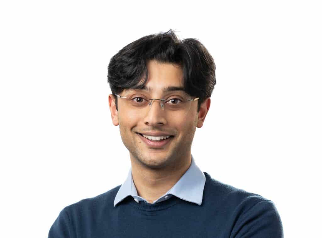 A headshot of Dr Roshan Karri. He has medium length black hir and is wearing glasses and a blue jumper over a blue collared shirt