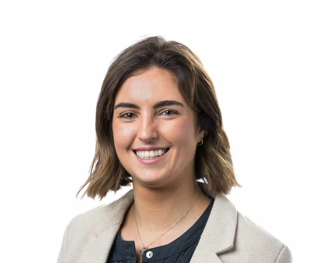 A headshot of Talia Clohessy. She has shoulder length dark hair, and is wearing a tan blazer over a black shirt.